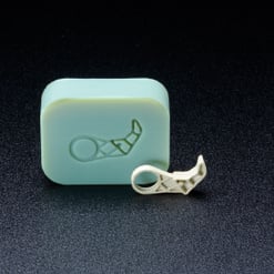  Das Bild zeigt ein hellgrünes, rechteckiges Formteil mit einer eingeprägten Form. Davor liegt ein kleines, weißes, unregelmäßig geformtes Objekt mit filigraner Struktur. Beide liegen auf dunklem, strukturiertem Hintergrund.