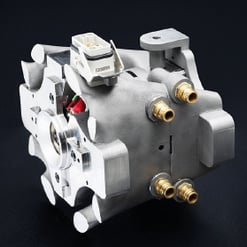  Das Bild zeigt ein komplexes, silbernes Bauteil mit verschiedenen Anschlüssen und einer Zahnrad-ähnlichen Struktur. Oben ist ein weißer Steckverbinder sichtbar.